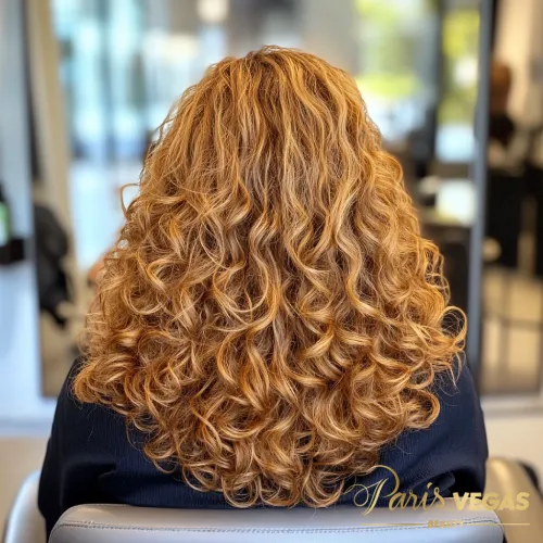 Mulher de costas com cabelo cacheado e loiro, em um ambiente de luxo com fundo desfocado.